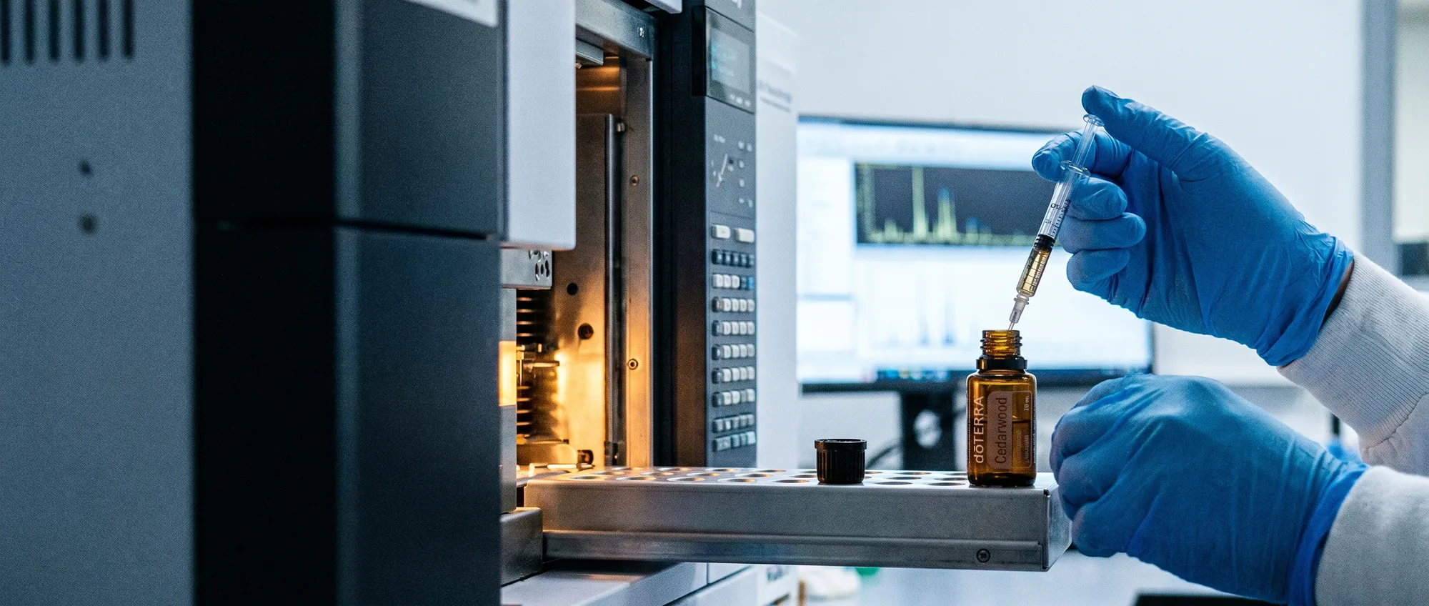 A scientist in nitrile gloves drawing a dōTERRA Cedarwood essential oil sample into a syringe for GC/MS analysis, a chromatogram on the monitor behind