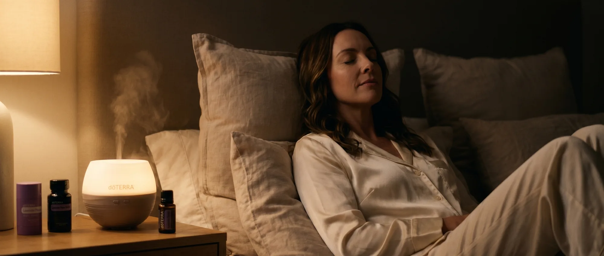 A woman resting calmly against linen pillows at night, eyes closed, as a dōTERRA Petal Diffuser mists beside Lavender Peace products on the nightstand