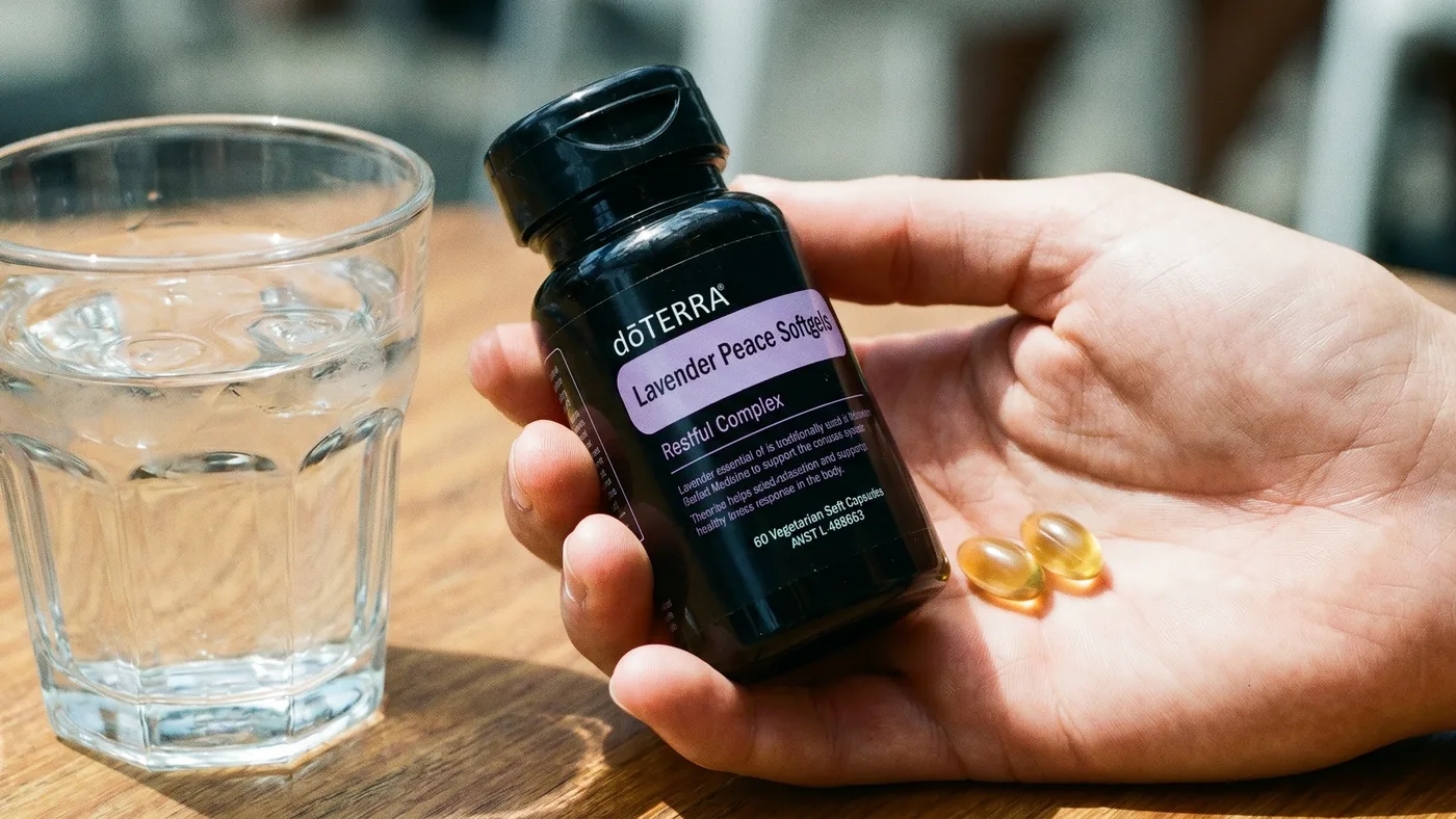 A hand holding the dōTERRA Lavender Peace Softgels bottle with two softgels in the palm and a glass of water