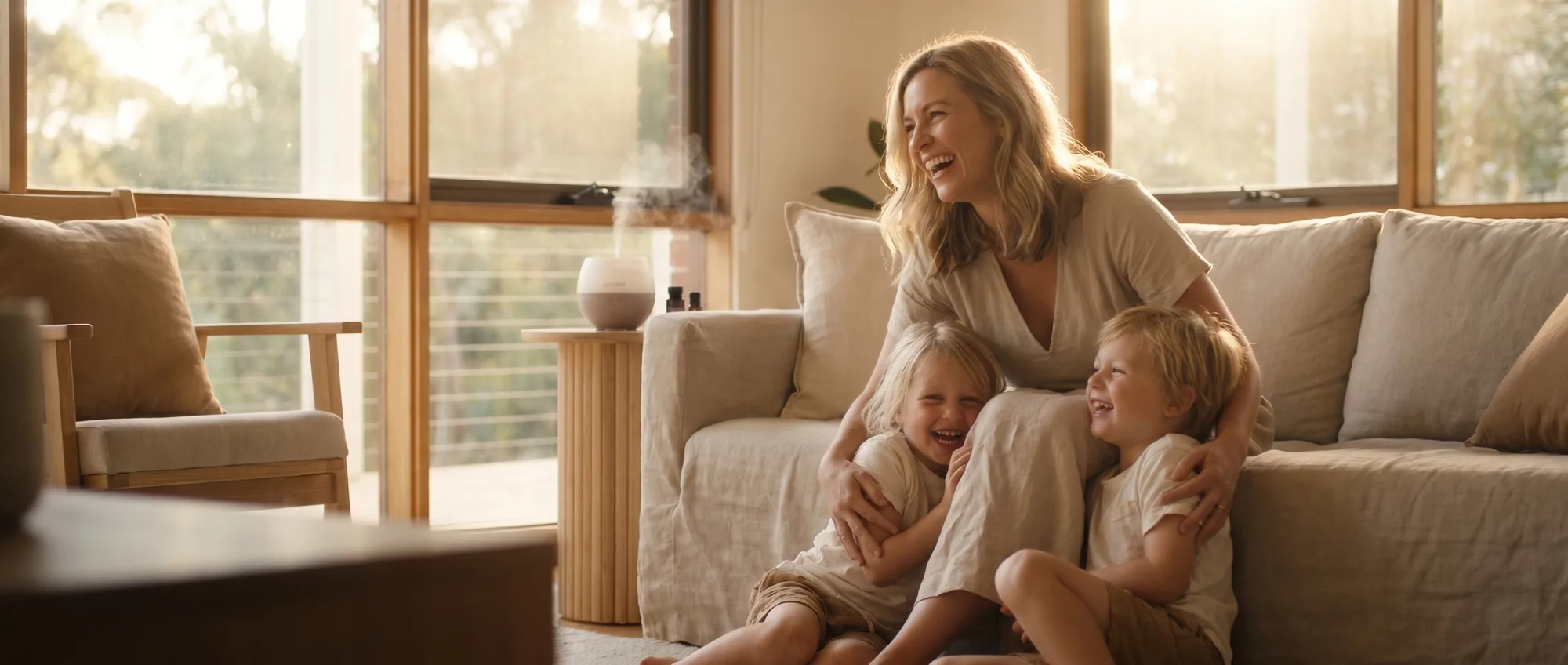 A mum laughing with her two young children in a warm, sunlit Australian living room, a dōTERRA Petal Diffuser misting on the side table