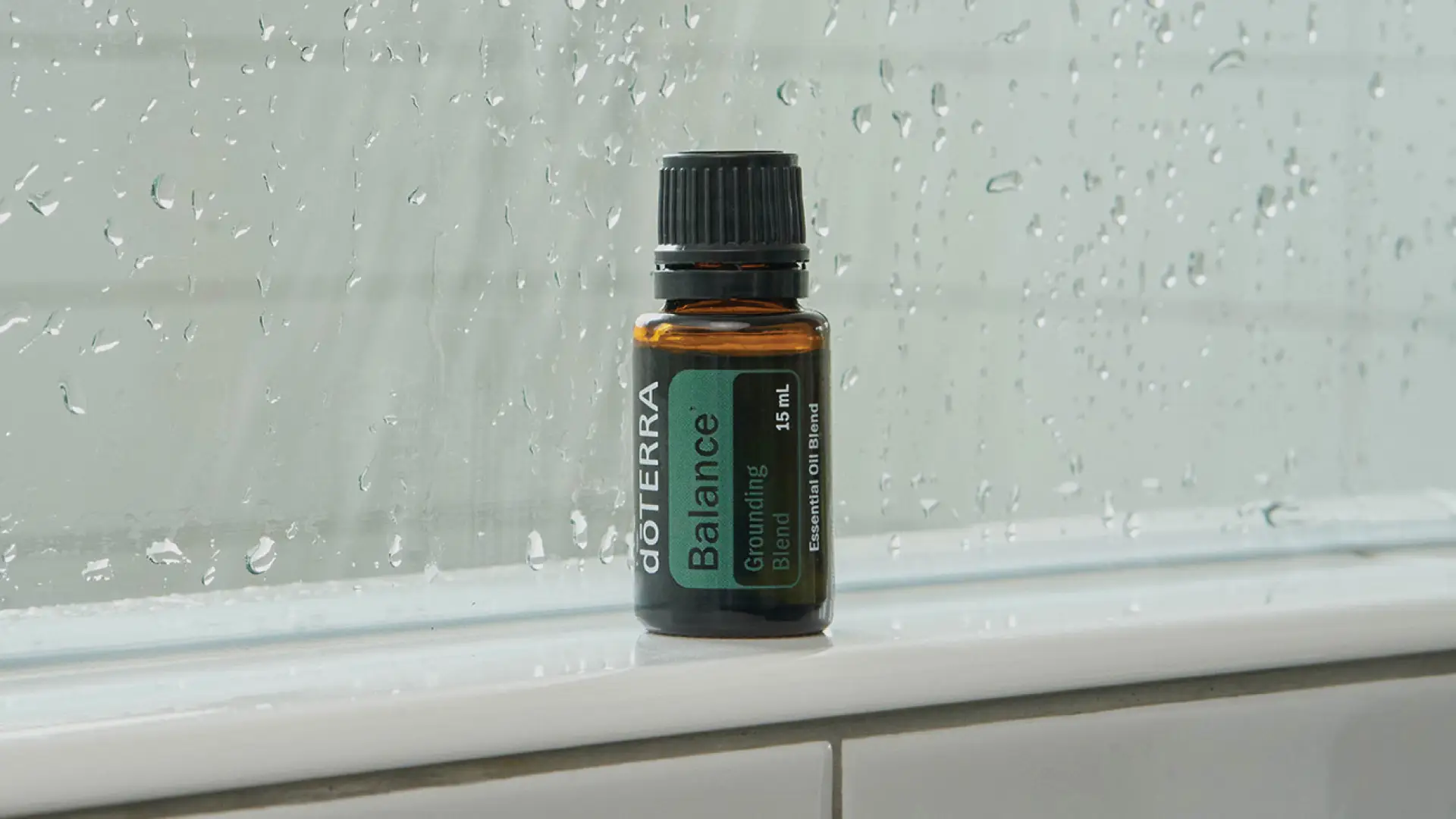 dōTERRA Balance Oil bottle placed on a wet bathroom windowsill with raindrops on the glass.