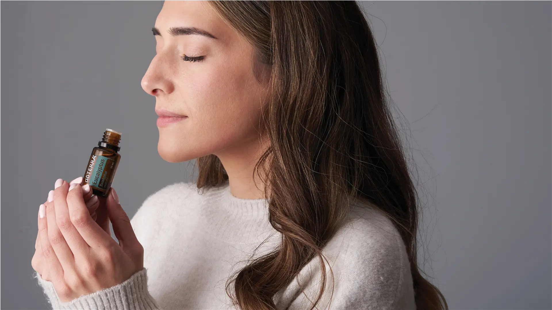 A woman with long brown hair and a soft white sweater gently holds a bottle of dōTERRA Eucalyptus Oil close to her nose, inhaling its refreshing aroma with a relaxed expression against a neutral grey background.