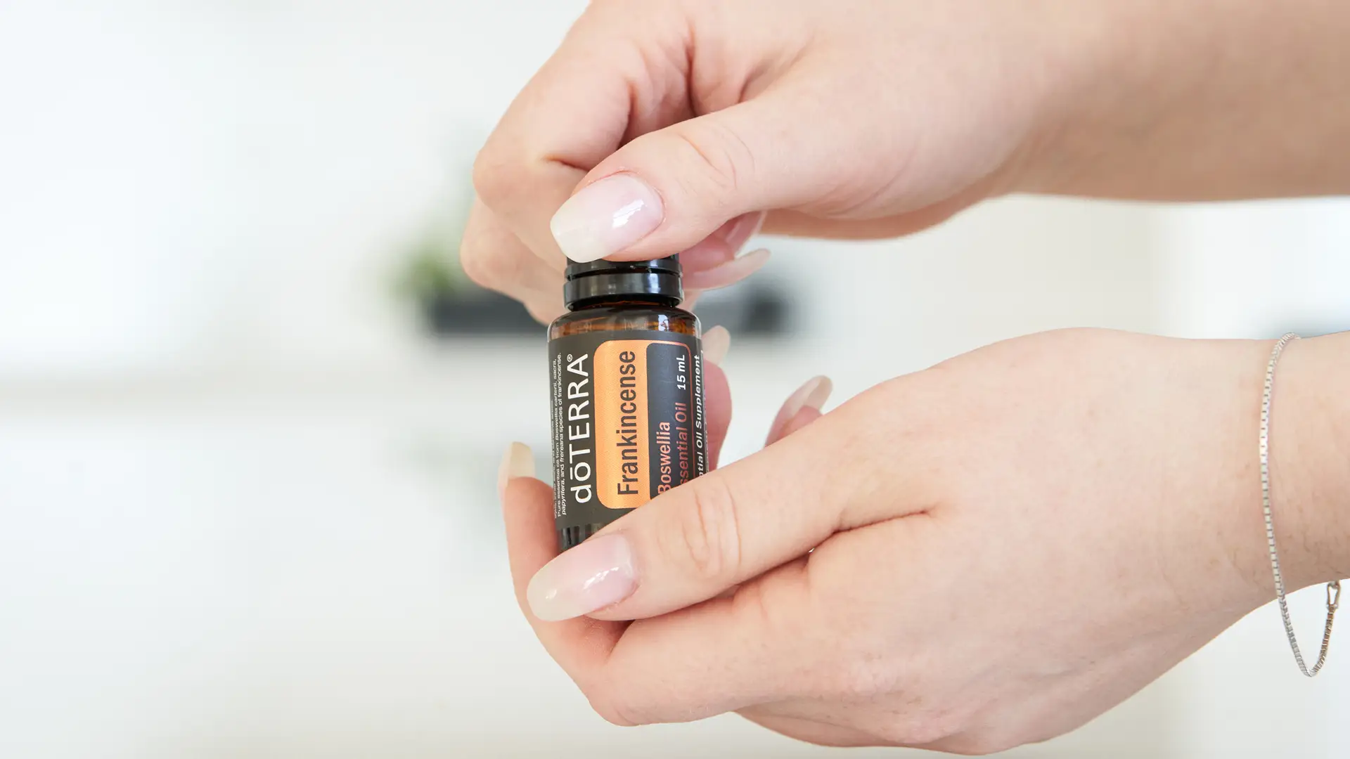 A person holding a 15 mL bottle of dōTERRA Frankincense Oil with neatly manicured nails, twisting the cap open in a bright, clean setting.