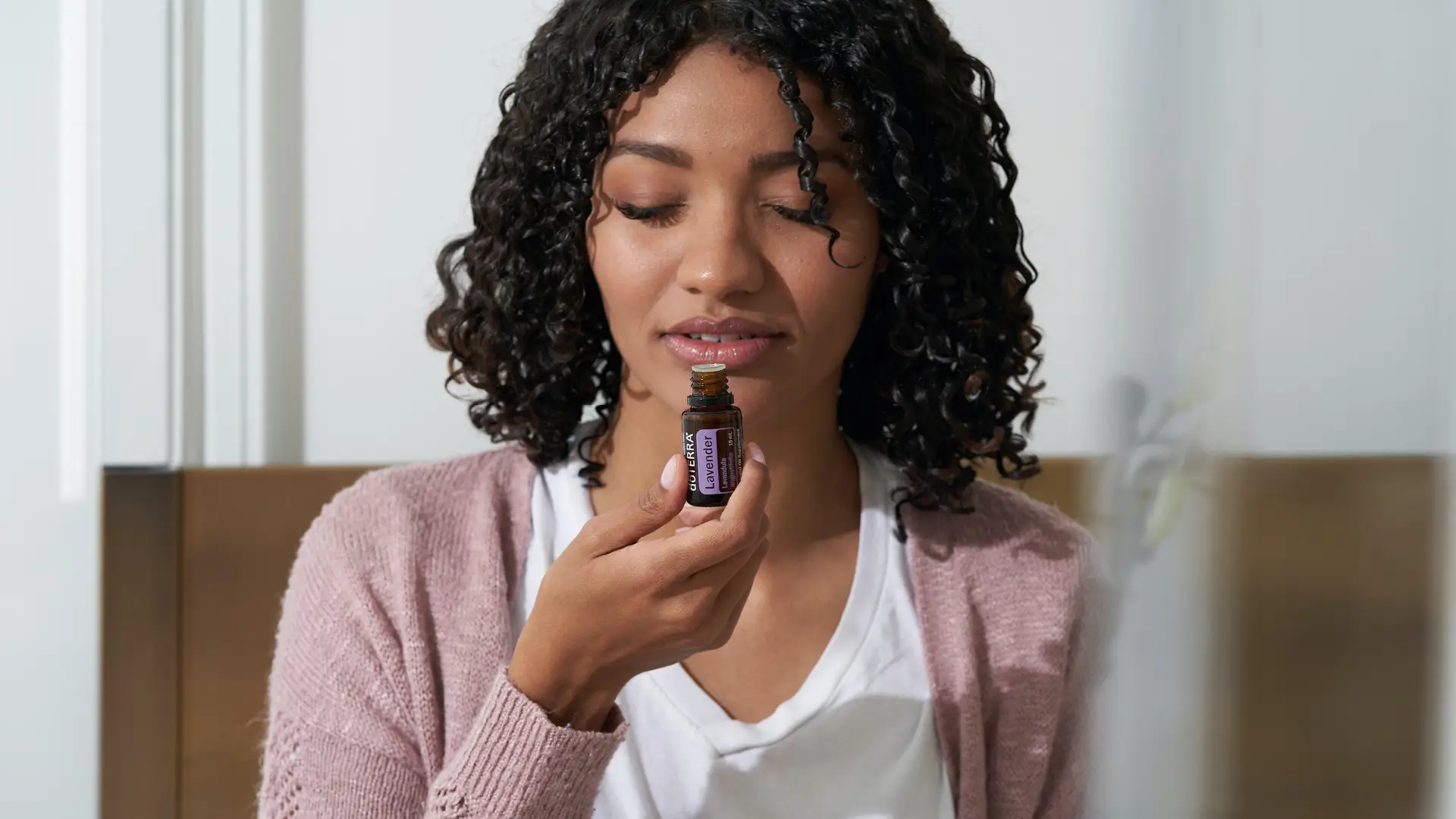 A woman with curly hair, wearing a pink cardigan, holds an open bottle of dōTERRA Lavender Oil close to her nose with a relaxed expression.