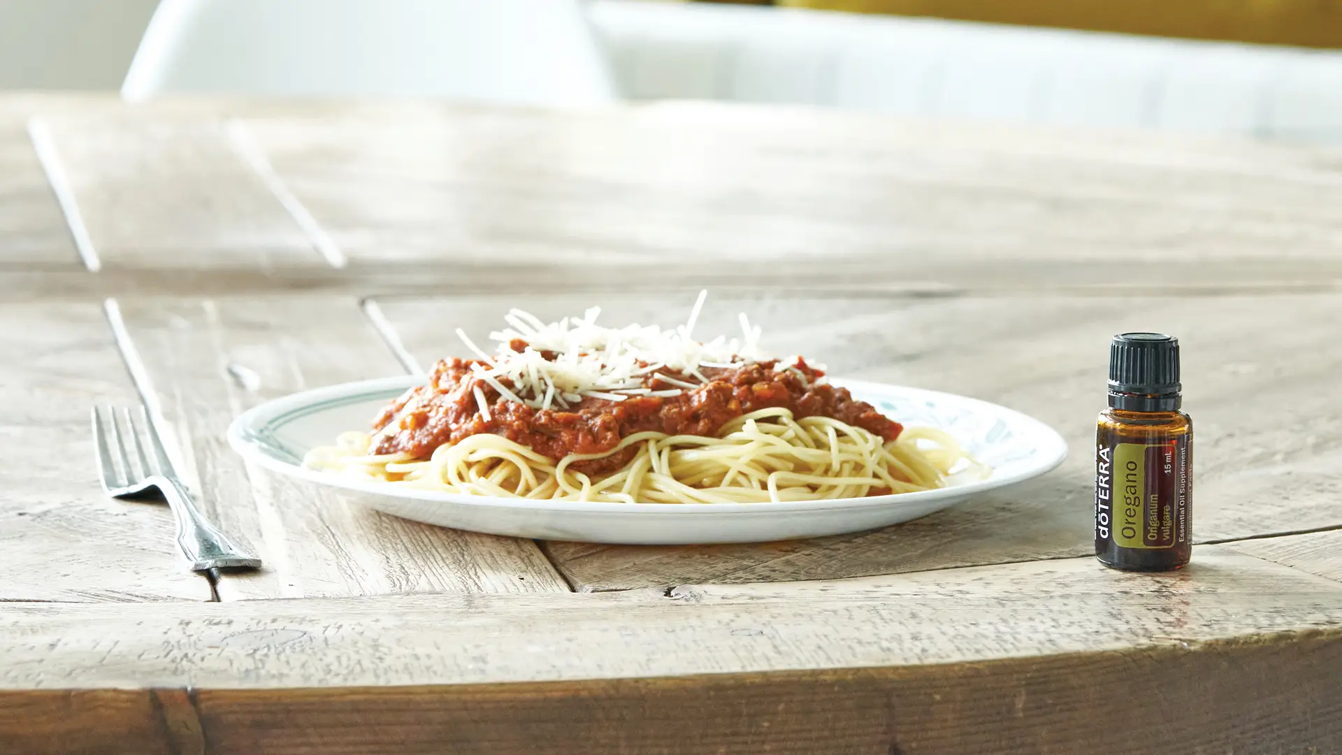 A bottle of dōTERRA Oregano Oil sits on a rustic wooden table next to a plate of spaghetti with rich tomato sauce and grated cheese.