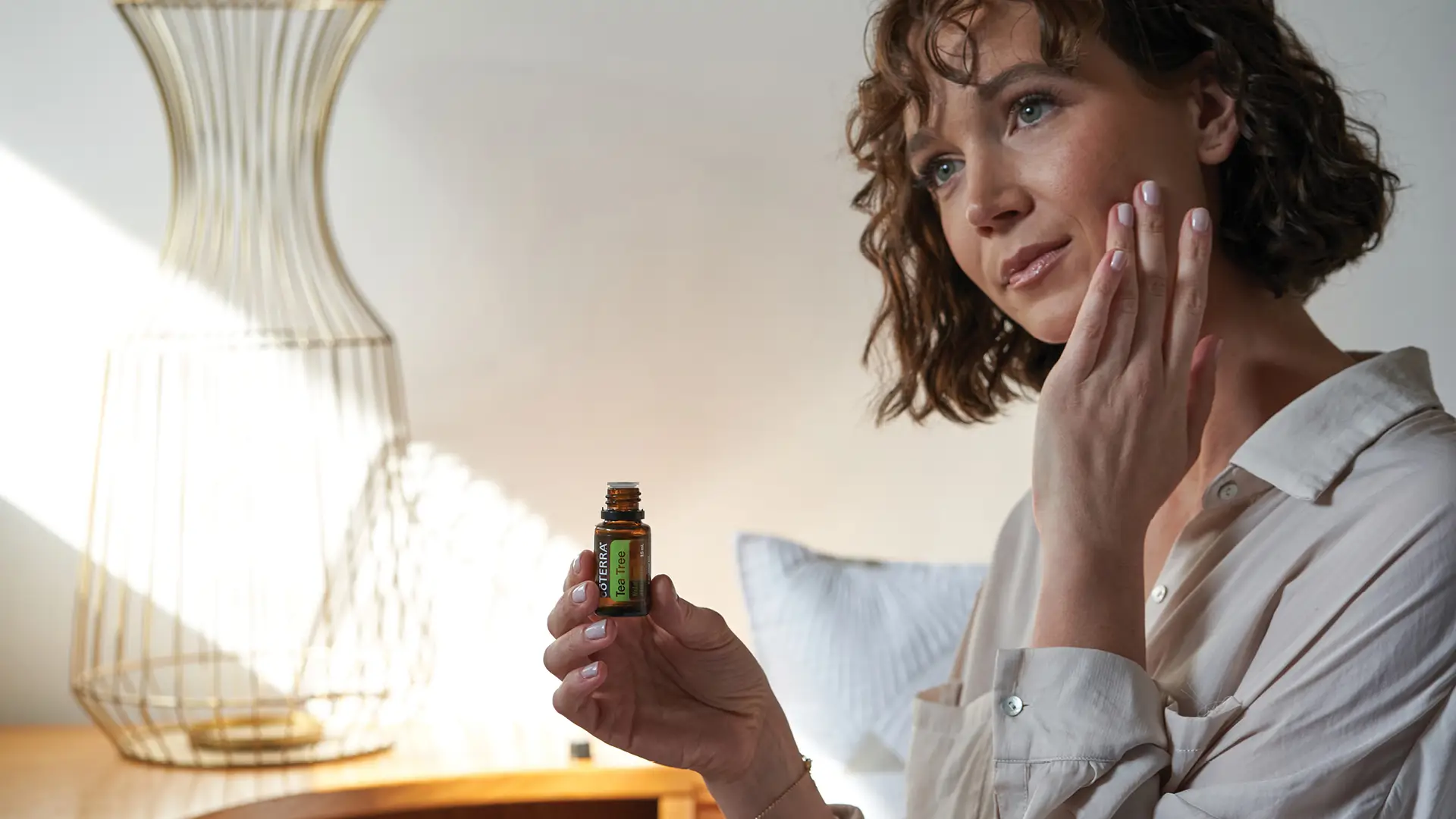 Woman applying dōTERRA Tea Tree Oil to her face with a gentle touch, holding the 15 mL essential oil bottle.