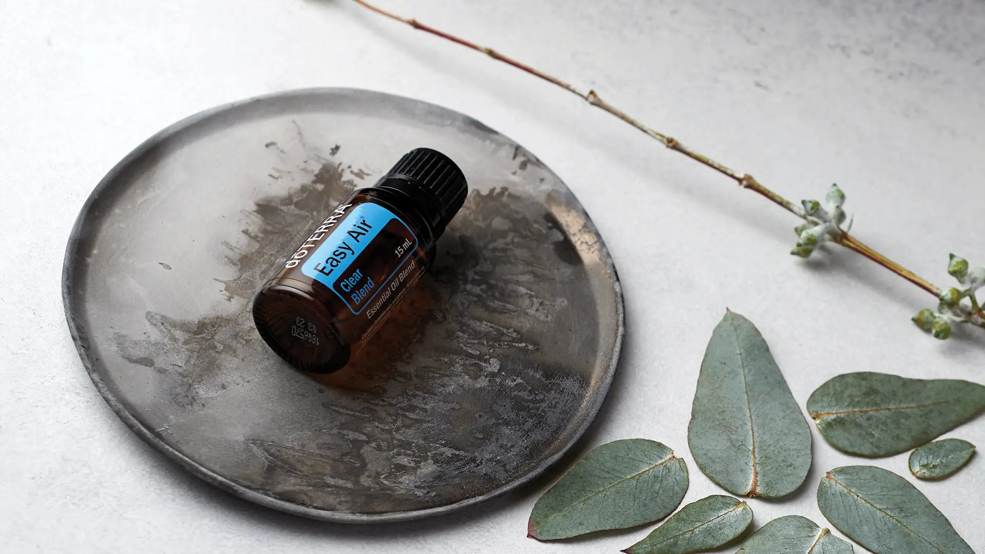 Brown glass bottle of dōTERRA Easy Air Blend resting on a grey ceramic plate, surrounded by eucalyptus leaves and a natural twig.