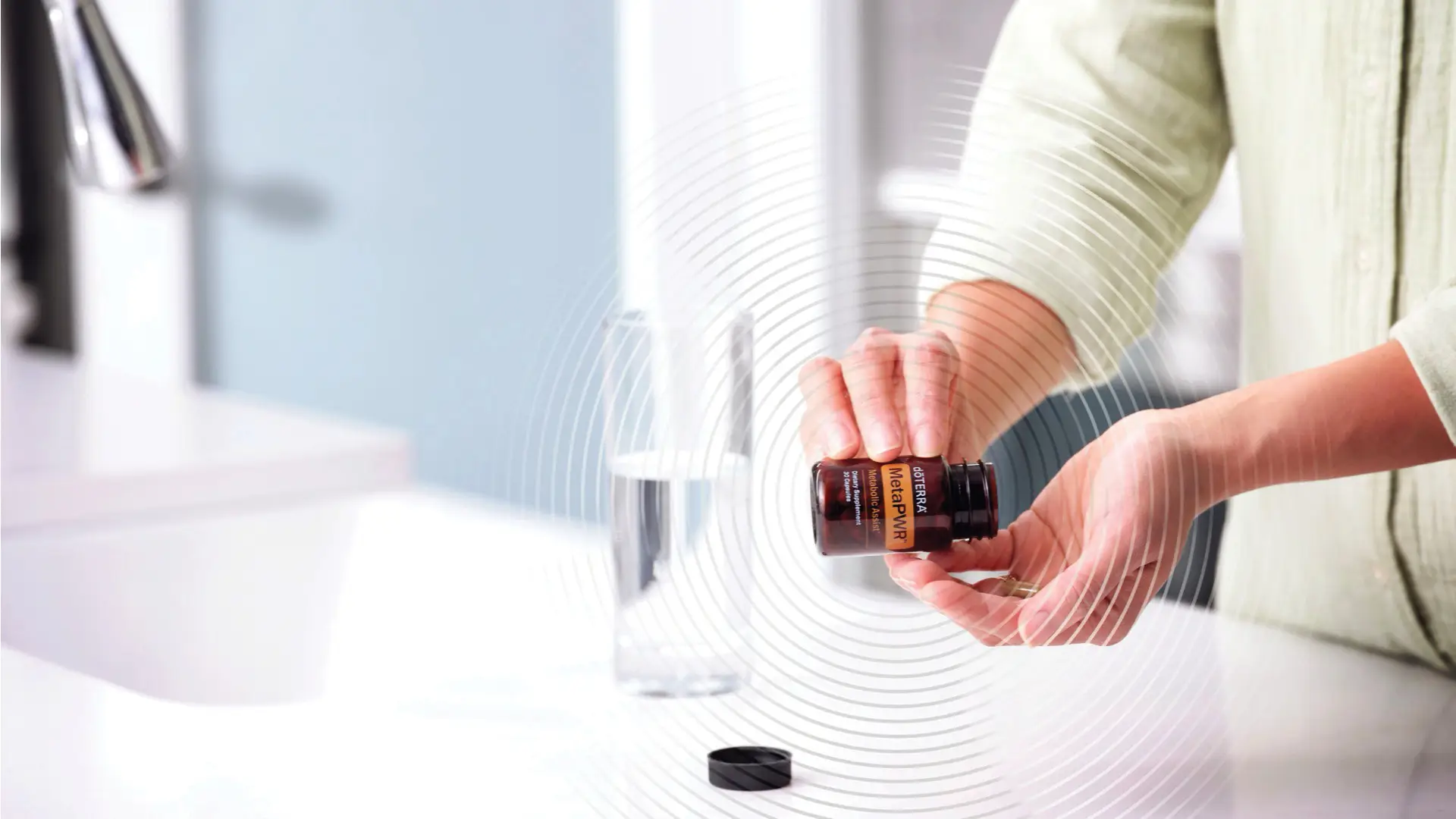 A person pours a capsule from a bottle of dōTERRA MetaPWR Assist beside a glass of water on a white bathroom counter.