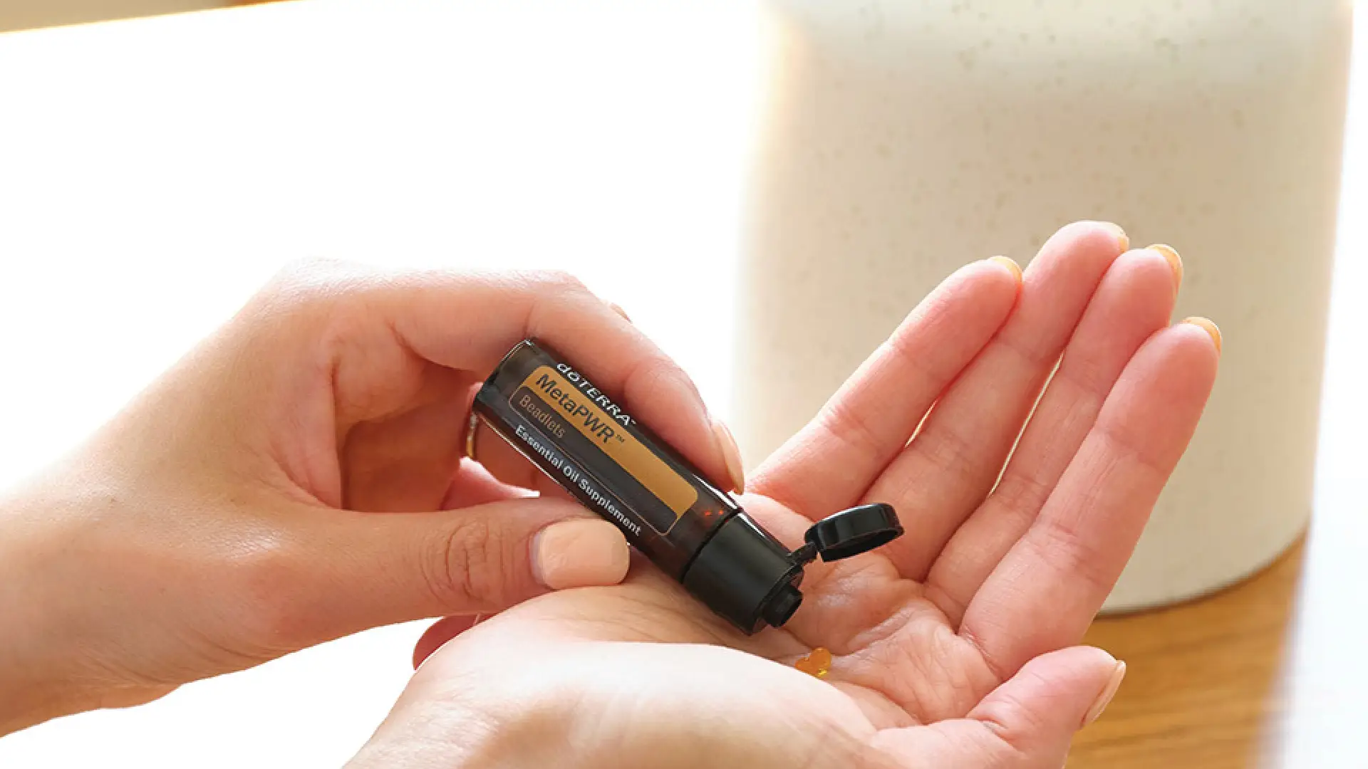 A person dispensing dōTERRA MetaPWR Flavour Beadlets into their hand from a small amber bottle, with soft natural light and a calm setting.