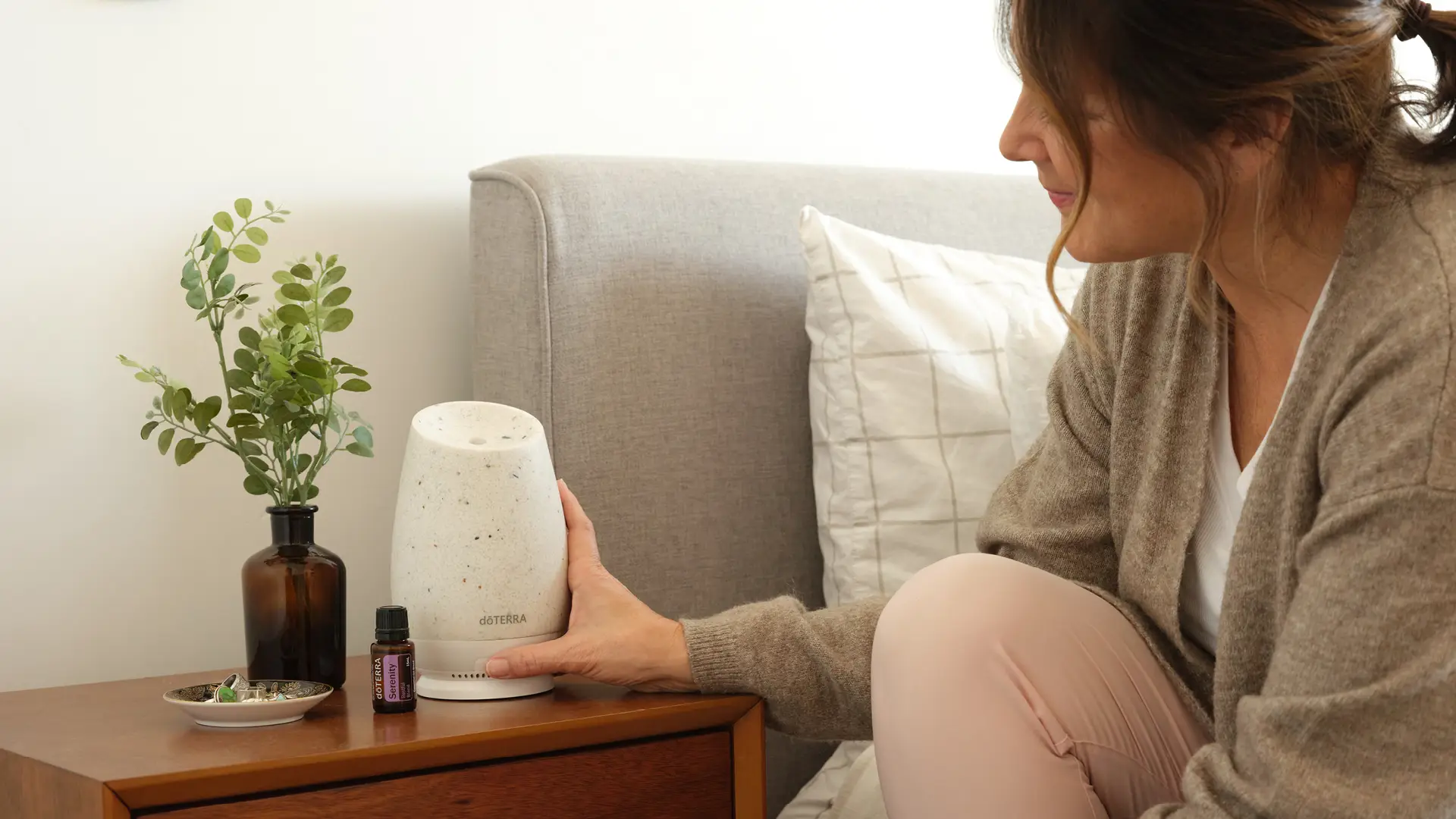 A woman preparing a diffuser on her bedside table next to a bottle of **dōTERRA Serenity Oil**, creating a calming nighttime routine.