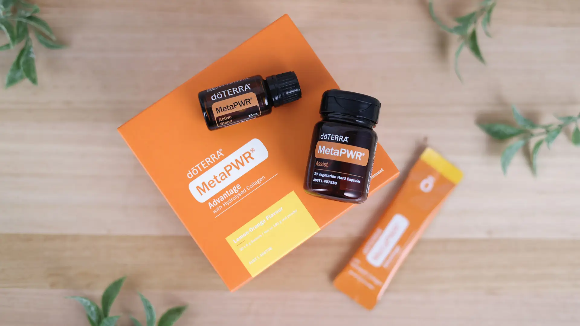 Flat lay of the dōTERRA MetaPWR Complete set featuring MetaPWR Advantage with Hydrolysed Collagen, MetaPWR Assist capsules, and MetaPWR Active Blend essential oil on a wooden surface with green leaves.